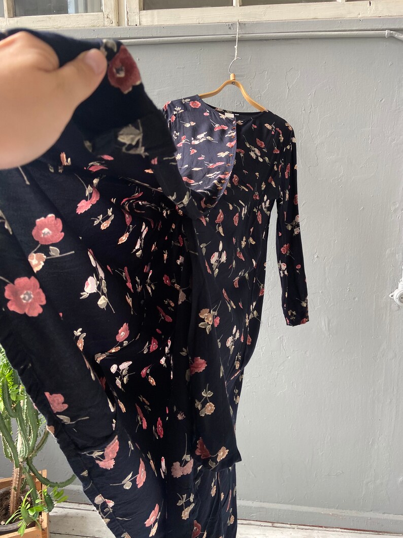 K&ouml;nnte beinhalten: Ein schwarzes Maxikleid mit Blumenmuster mit rosa und wei&szlig;en Bl&uuml;ten. Das Kleid hat lange &Auml;rmel und einen Knopfleisten vorne.