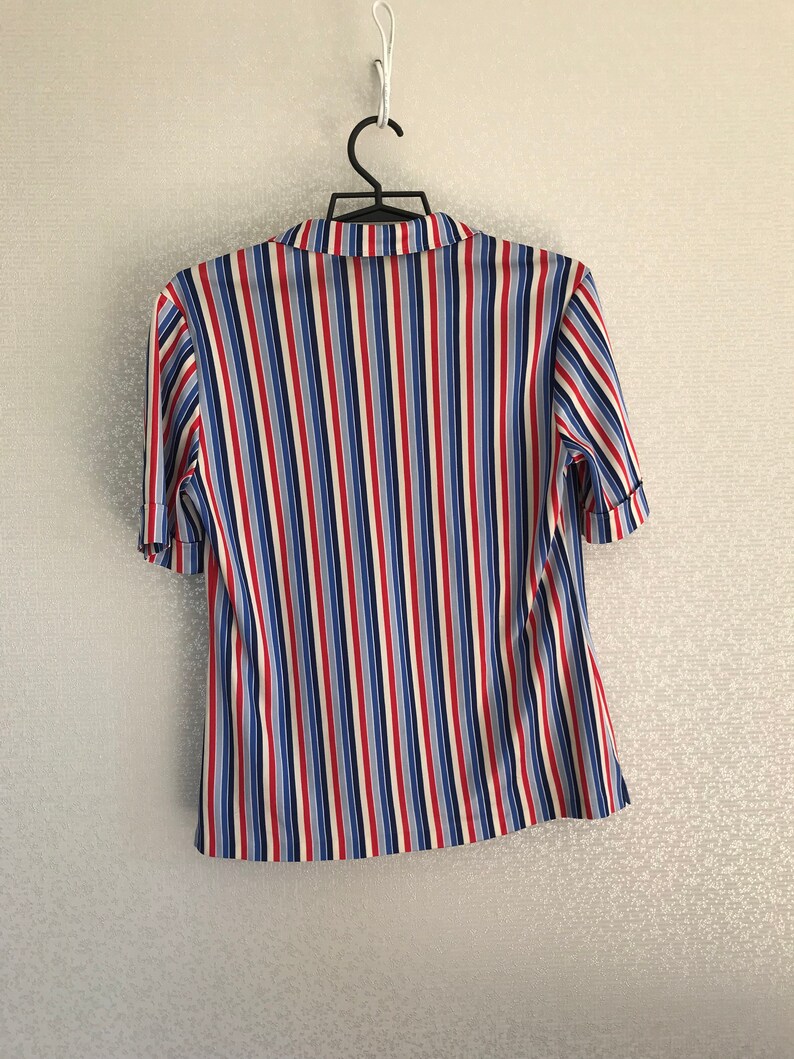 May include: A striped short-sleeved shirt with red, blue, and white vertical stripes. The shirt has a button-up collar and is hanging on a hanger.