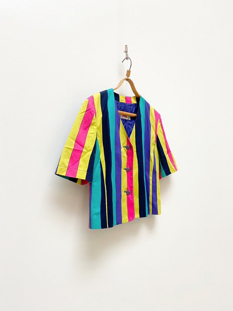 May include: A short-sleeved, multi-coloured striped blazer with a V-neck and button closure. The blazer is hanging on a wooden hanger against a white background.