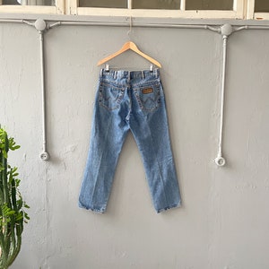 Pu&ograve; includere: Un paio di jeans in denim blu chiaro appesi a un appendiabiti in legno contro un muro grigio. I jeans hanno una toppa in pelle marrone sul retro con il logo Wrangler.
