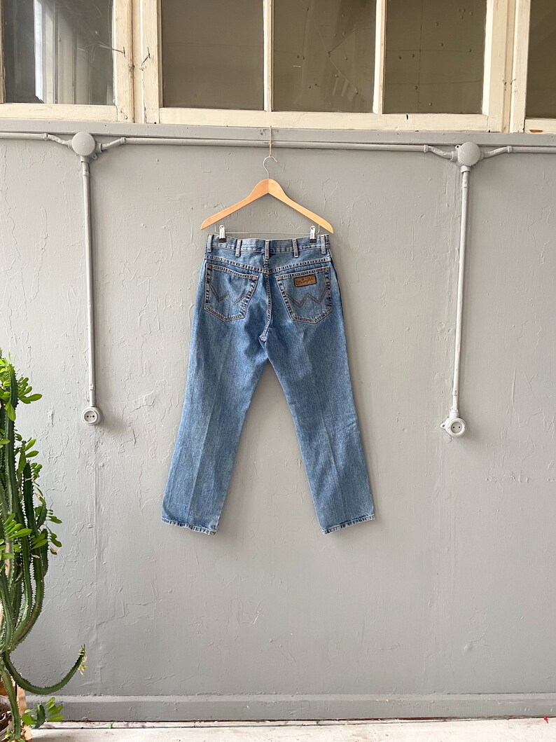 Pu&ograve; includere: Un paio di jeans in denim blu chiaro appesi a un appendiabiti in legno contro un muro grigio. I jeans hanno una toppa in pelle marrone sul retro con il logo Wrangler.
