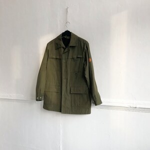 May include: A green utility jacket with a patch on the right sleeve and a button closure.