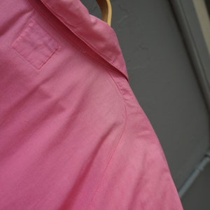 May include: A pink button-down shirt with a single pocket on the left side of the chest. The shirt is hanging on a wooden hanger.