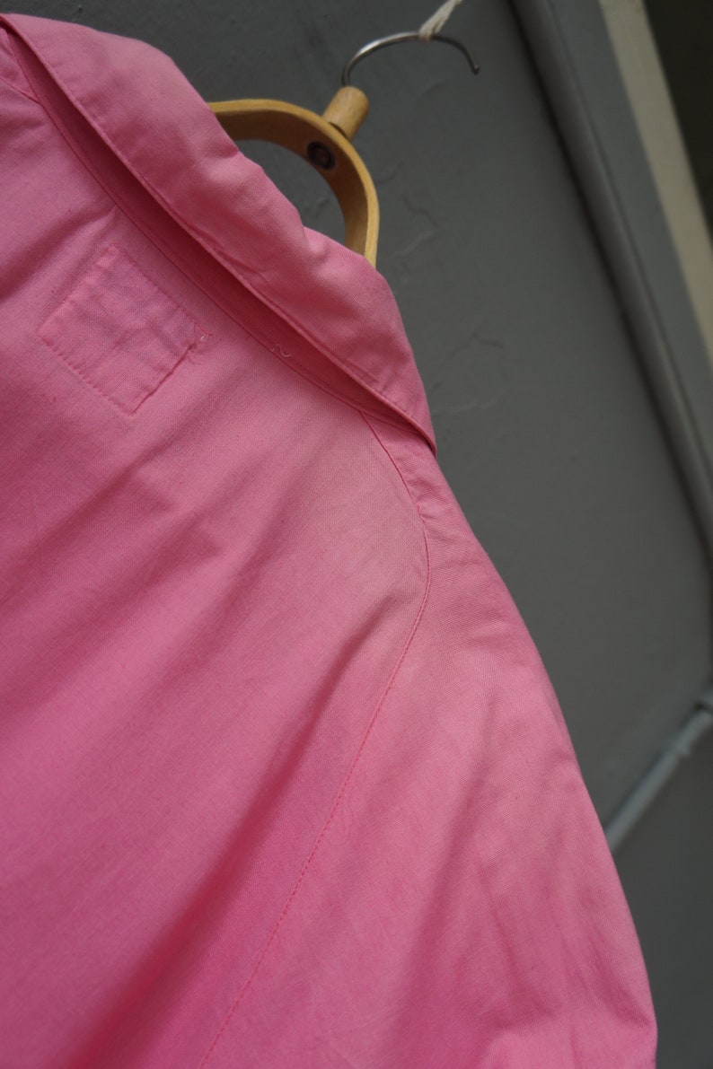 May include: A pink button-down shirt with a single pocket on the left side of the chest. The shirt is hanging on a wooden hanger.