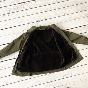 May include: A green jacket with a brown fleece lining. The jacket is unzipped and laid flat on a white wooden surface.