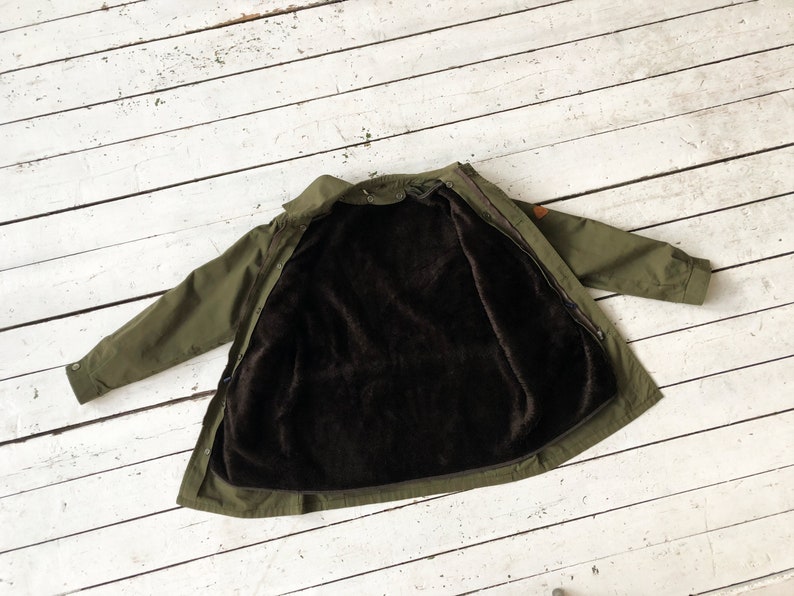May include: A green jacket with a brown fleece lining. The jacket is unzipped and laid flat on a white wooden surface.