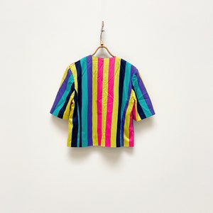 May include: A short-sleeved, multi-coloured striped blouse with a yellow, pink, blue, purple, and black colour scheme. The blouse is hanging on a wooden hanger against a white background.
