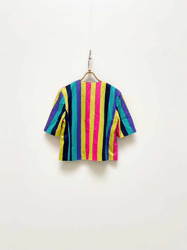 May include: A short-sleeved, multi-coloured striped blouse with a yellow, pink, blue, purple, and black colour scheme. The blouse is hanging on a wooden hanger against a white background.