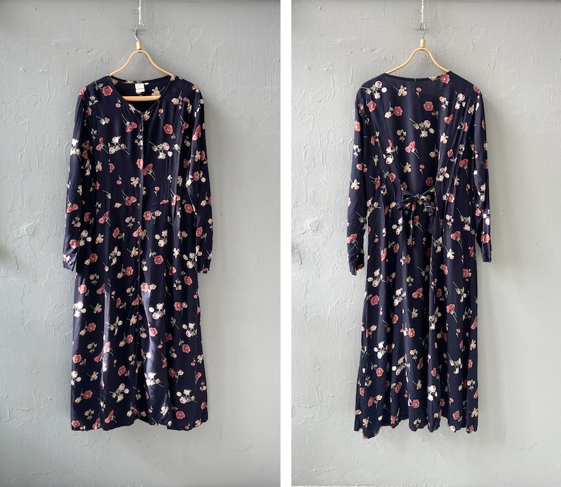 K&ouml;nnte beinhalten: Ein lang&auml;rmeliges Blumenkleid mit dunkelblauem Hintergrund und rosa und wei&szlig;en Blumen. Das Kleid hat einen V-Ausschnitt und einen Knopfleisten vorne. Das Kleid ist aus einem leichten Stoff gefertigt und hat eine flie&szlig;ende Silhouette.