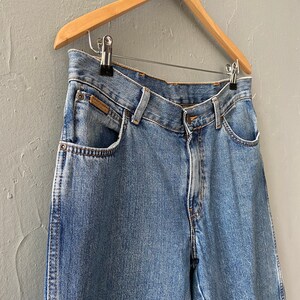 Pu&ograve; includere: Jeans in denim blu chiaro con un classico stile a cinque tasche. I jeans sono appesi a un appendiabiti in legno.