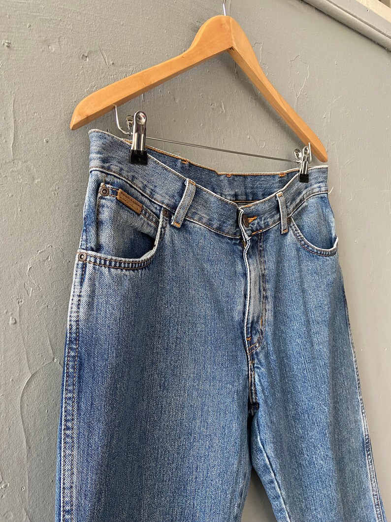 Pu&ograve; includere: Jeans in denim blu chiaro con un classico stile a cinque tasche. I jeans sono appesi a un appendiabiti in legno.