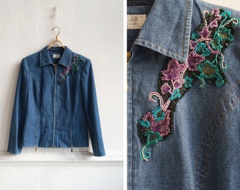 以下が含まれることがあります： 右肩に花の刺繍が施された青いデニムジャケット。刺繍には紫、ターコイズ、黒のビーズが使われています。