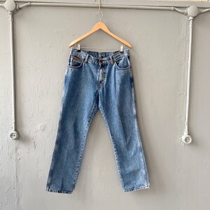 Pu&ograve; includere: Un paio di jeans in denim blu chiaro appesi a un appendiabiti in legno contro un muro grigio.
