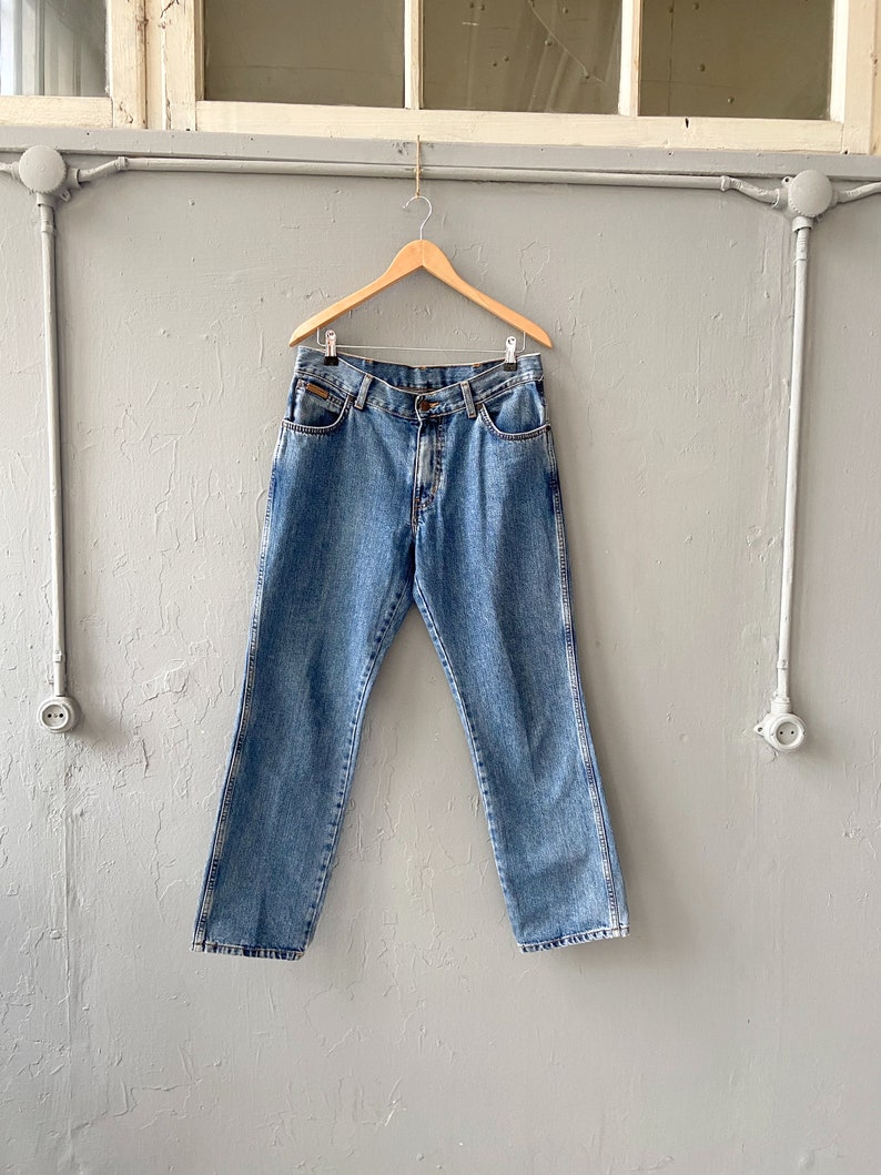 Pu&ograve; includere: Un paio di jeans in denim blu chiaro appesi a un appendiabiti in legno contro un muro grigio.