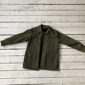 May include: A green utility jacket with a button closure, two large pockets, and a small patch on the right sleeve.