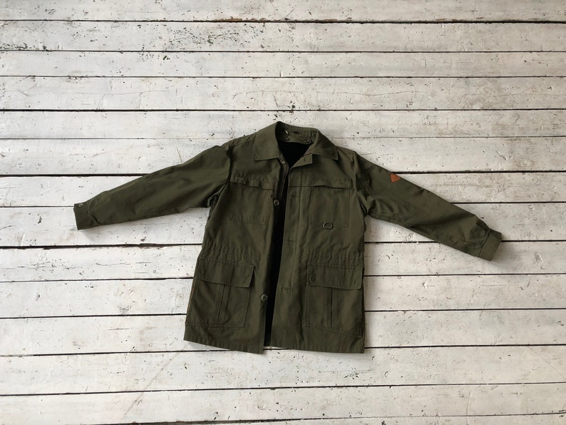 May include: A green utility jacket with a button closure, two large pockets, and a small patch on the right sleeve.