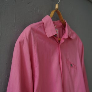 May include: A pink long-sleeved button-down shirt hanging on a wooden hanger. The shirt has a single pocket on the left side and a button-up collar.