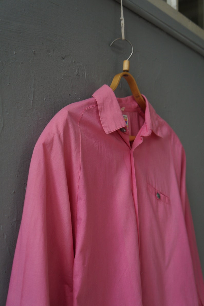 May include: A pink long-sleeved button-down shirt hanging on a wooden hanger. The shirt has a single pocket on the left side and a button-up collar.