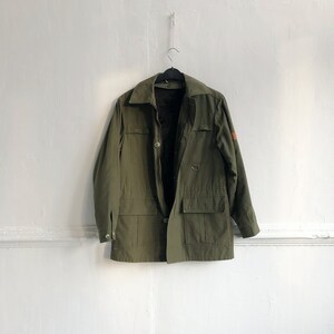 May include: A green utility jacket with a brown patch on the sleeve. The jacket has a button closure and multiple pockets.