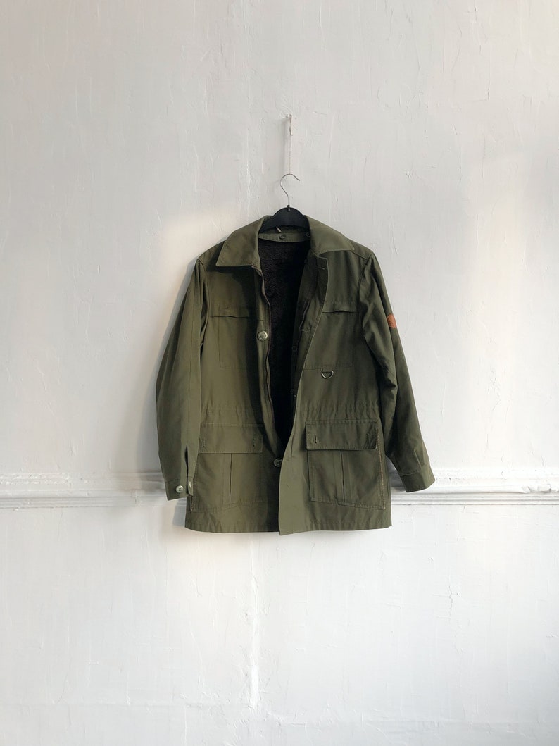 May include: A green utility jacket with a brown patch on the sleeve. The jacket has a button closure and multiple pockets.