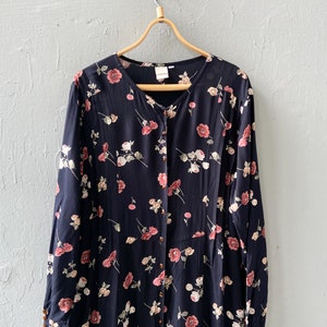 K&ouml;nnte beinhalten: Ein lang&auml;rmeliges schwarzes Kleid mit einem Blumendruck mit rosa und wei&szlig;en Blumen. Das Kleid hat einen Knopfleisten vorne und einen V-Ausschnitt.