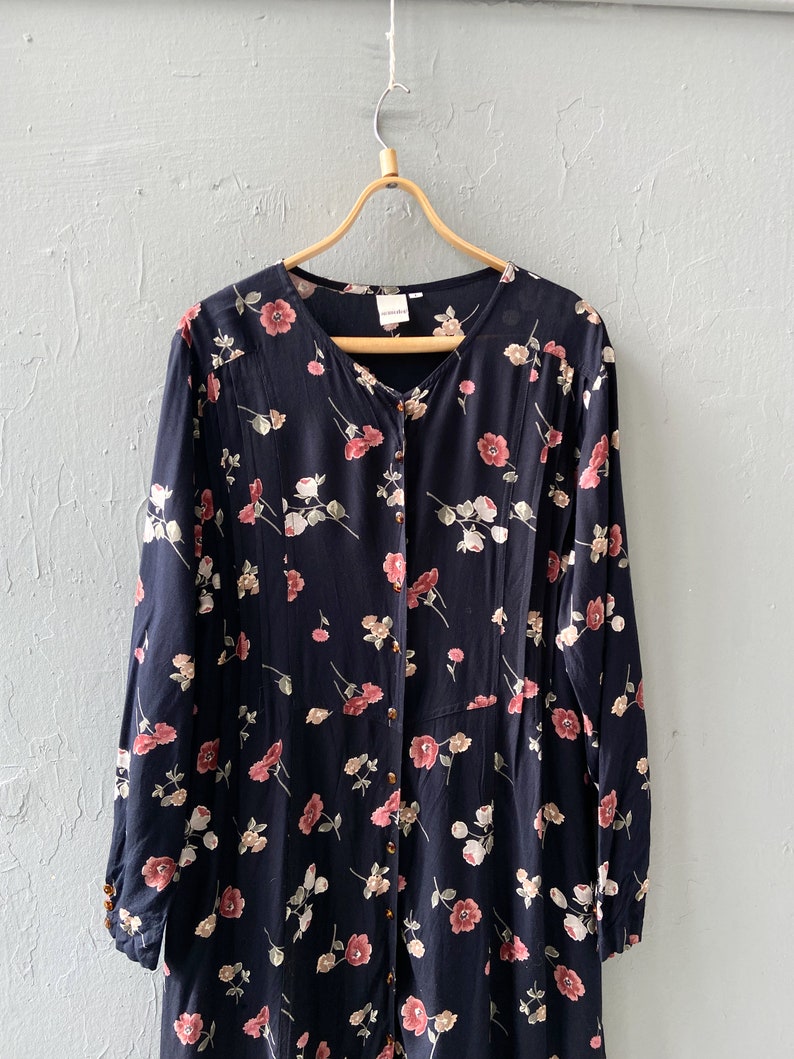 K&ouml;nnte beinhalten: Ein lang&auml;rmeliges schwarzes Kleid mit einem Blumendruck mit rosa und wei&szlig;en Blumen. Das Kleid hat einen Knopfleisten vorne und einen V-Ausschnitt.
