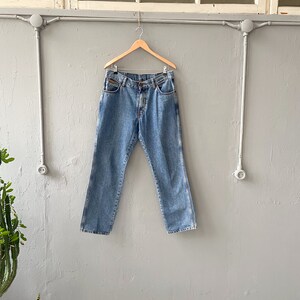 Pu&ograve; includere: Un paio di jeans in denim blu chiaro appesi a un appendiabiti in legno contro un muro grigio.