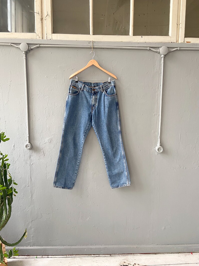 Pu&ograve; includere: Un paio di jeans in denim blu chiaro appesi a un appendiabiti in legno contro un muro grigio.