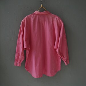May include: A pink long-sleeved button-up shirt hanging on a wooden hanger against a gray wall.