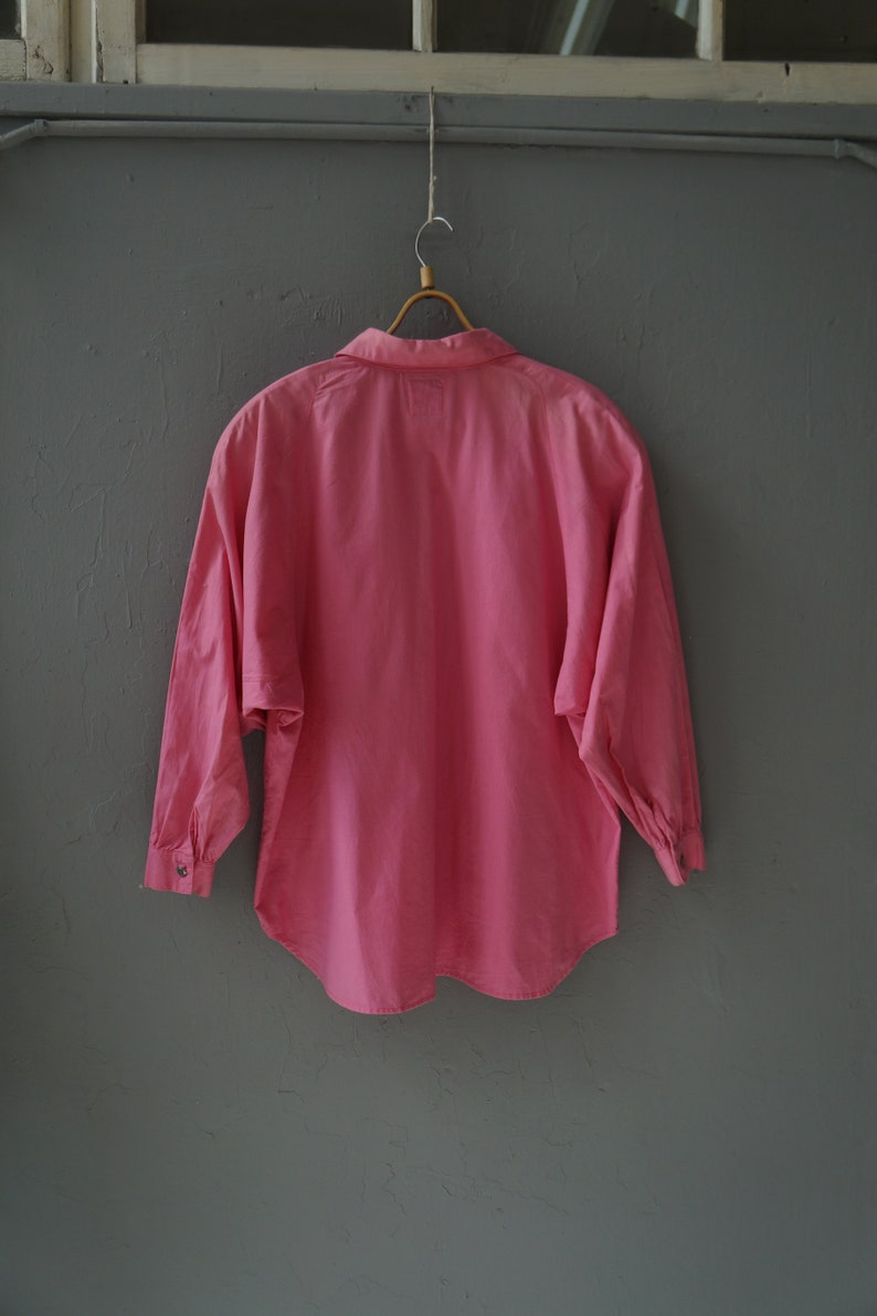May include: A pink long-sleeved button-up shirt hanging on a wooden hanger against a gray wall.