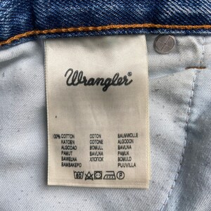 Pu&ograve; includere: Un'etichetta di cura bianca con testo nero che recita "Wrangler" e elenca la composizione del tessuto come 100% cotone in pi&ugrave; lingue. L'etichetta include anche istruzioni di cura con simboli per il lavaggio in lavatrice, la candeggina, l'asciugatura in asciugatrice, il ferro da stiro e la pulizia a secco.