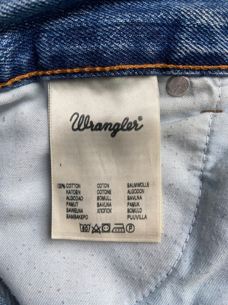 Pu&ograve; includere: Un'etichetta di cura bianca con testo nero che recita "Wrangler" e elenca la composizione del tessuto come 100% cotone in pi&ugrave; lingue. L'etichetta include anche istruzioni di cura con simboli per il lavaggio in lavatrice, la candeggina, l'asciugatura in asciugatrice, il ferro da stiro e la pulizia a secco.