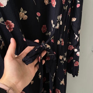 K&ouml;nnte beinhalten: Ein schwarzes Maxikleid mit Blumenmuster in Rosa und Wei&szlig;. Das Kleid hat einen G&uuml;rtel zum Binden in der Taille und lange &Auml;rmel.