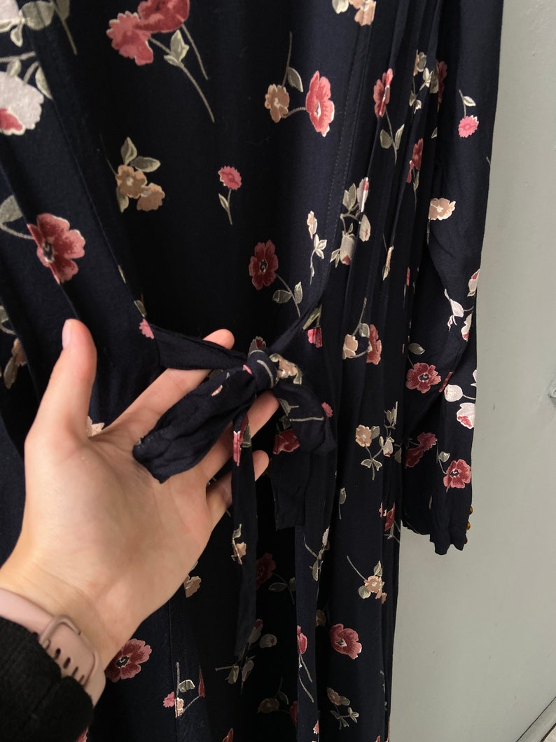 K&ouml;nnte beinhalten: Ein schwarzes Maxikleid mit Blumenmuster in Rosa und Wei&szlig;. Das Kleid hat einen G&uuml;rtel zum Binden in der Taille und lange &Auml;rmel.