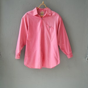 May include: A pink long-sleeved button-down shirt with a small pocket on the left side. The shirt has a label that says "Sun Wear" on it.