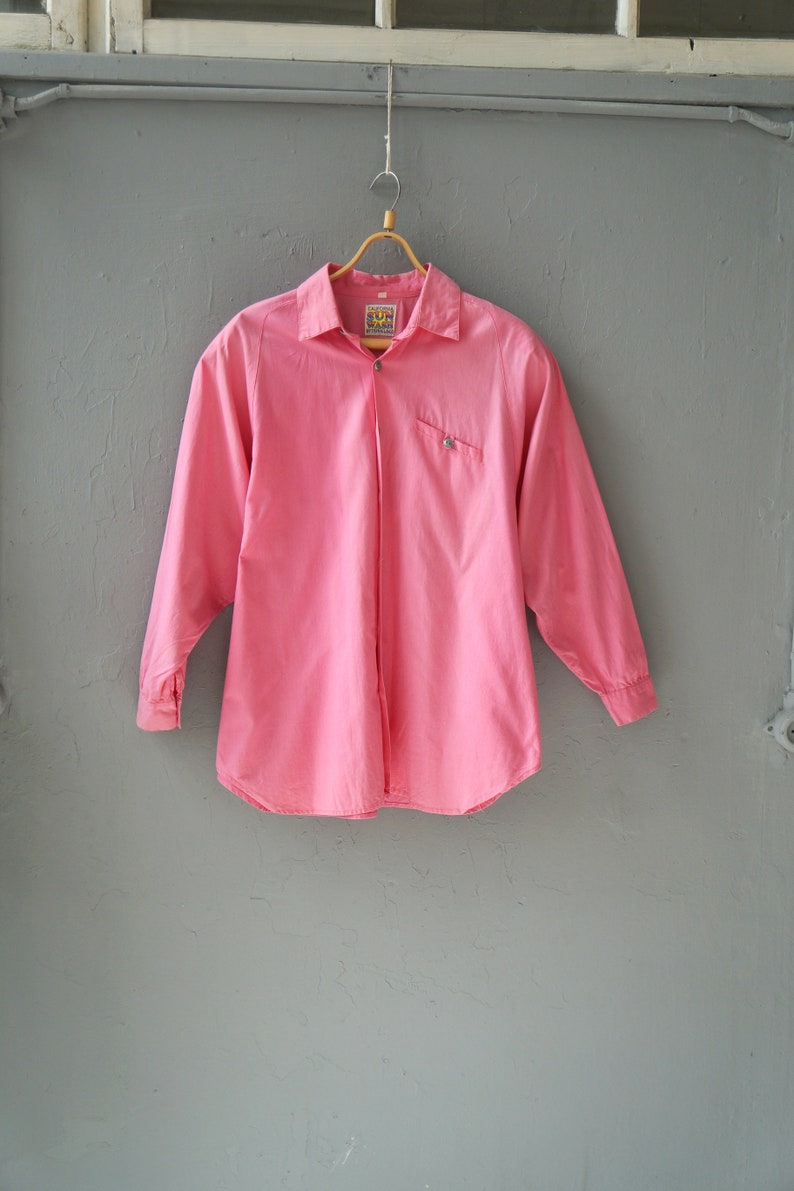 May include: A pink long-sleeved button-down shirt with a small pocket on the left side. The shirt has a label that says "Sun Wear" on it.