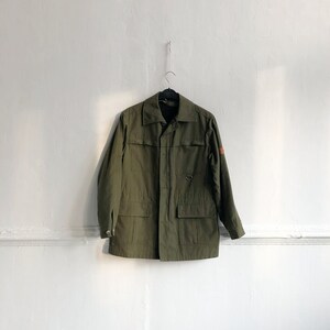 May include: A green utility jacket with a button closure, two large pockets, and a small patch on the right sleeve.