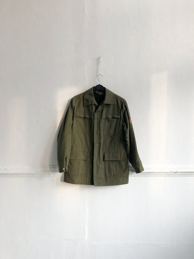 May include: A green utility jacket with a button closure, two large pockets, and a small patch on the right sleeve.