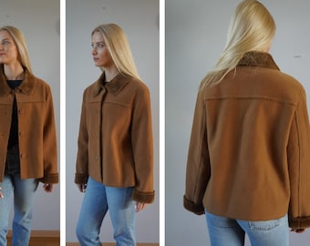 Könnte beinhalten: Eine braune Wildlederjacke mit einem Kunstfellkragen und -ärmelaufschlägen. Die Jacke hat einen Knopfverschluss und eine leicht verkürzte Länge.