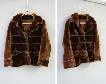 Puede incluir: Una chaqueta de piel sintética marrón con cierre de botones y dos bolsillos. La chaqueta tiene cuello y mangas largas.