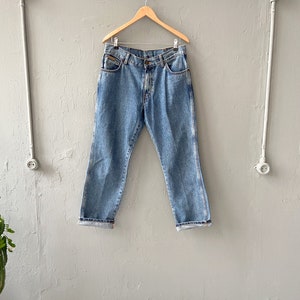 Pu&ograve; includere: Un paio di jeans in denim blu chiaro appesi a un appendiabiti in legno contro un muro grigio.