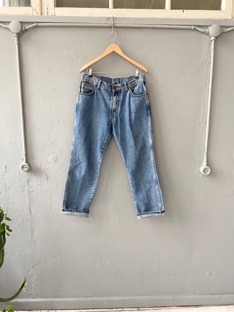 Pu&ograve; includere: Un paio di jeans in denim blu chiaro appesi a un appendiabiti in legno contro un muro grigio.