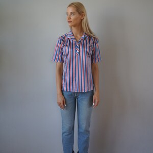 May include: A woman wearing a striped short-sleeved shirt with red, white, and blue stripes. She is also wearing blue jeans and white sneakers.