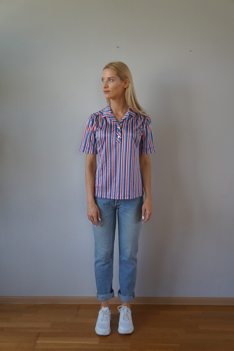 May include: A woman wearing a striped short-sleeved shirt with red, white, and blue stripes. She is also wearing blue jeans and white sneakers.