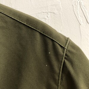 May include: Close-up of the shoulder seam of a green jacket. The fabric is a tightly woven twill.