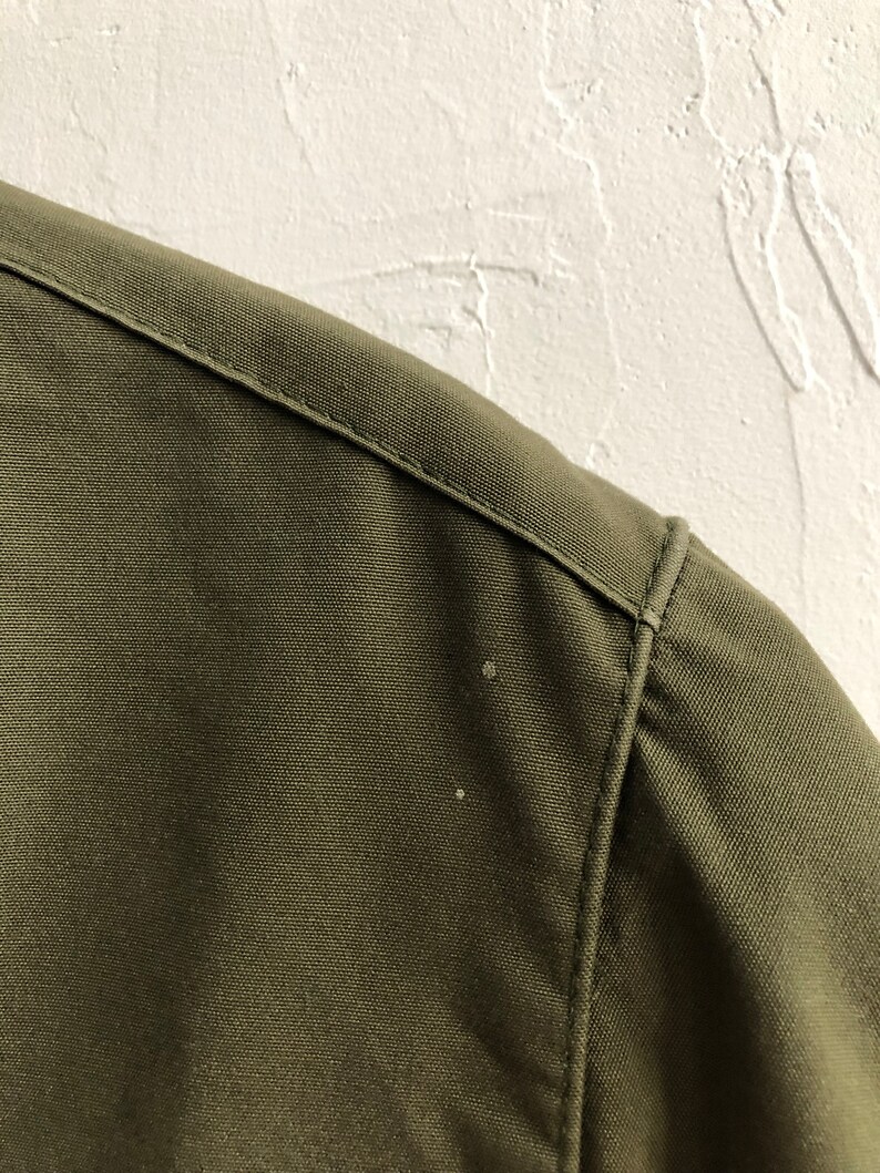 May include: Close-up of the shoulder seam of a green jacket. The fabric is a tightly woven twill.