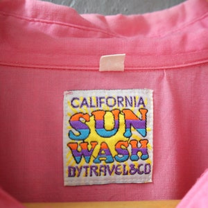 May include: A pink button-down shirt with a white label that says "CALIFORNIA SUN WASH BY TRAVEL & CO."