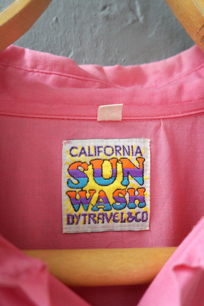 May include: A pink button-down shirt with a white label that says "CALIFORNIA SUN WASH BY TRAVEL & CO."