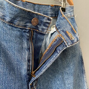 Pu&ograve; includere: Un paio di jeans blu denim con una zip e una toppa in pelle marrone con la scritta "Wrangler" su di essa.