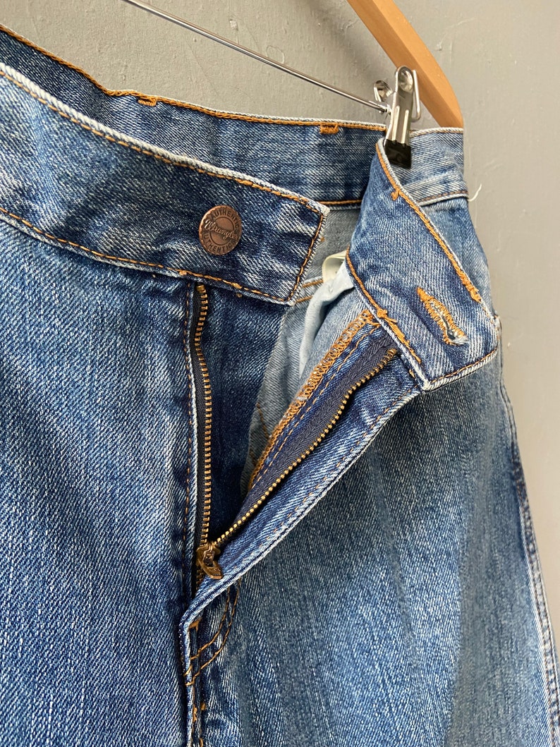 Pu&ograve; includere: Un paio di jeans blu denim con una zip e una toppa in pelle marrone con la scritta "Wrangler" su di essa.